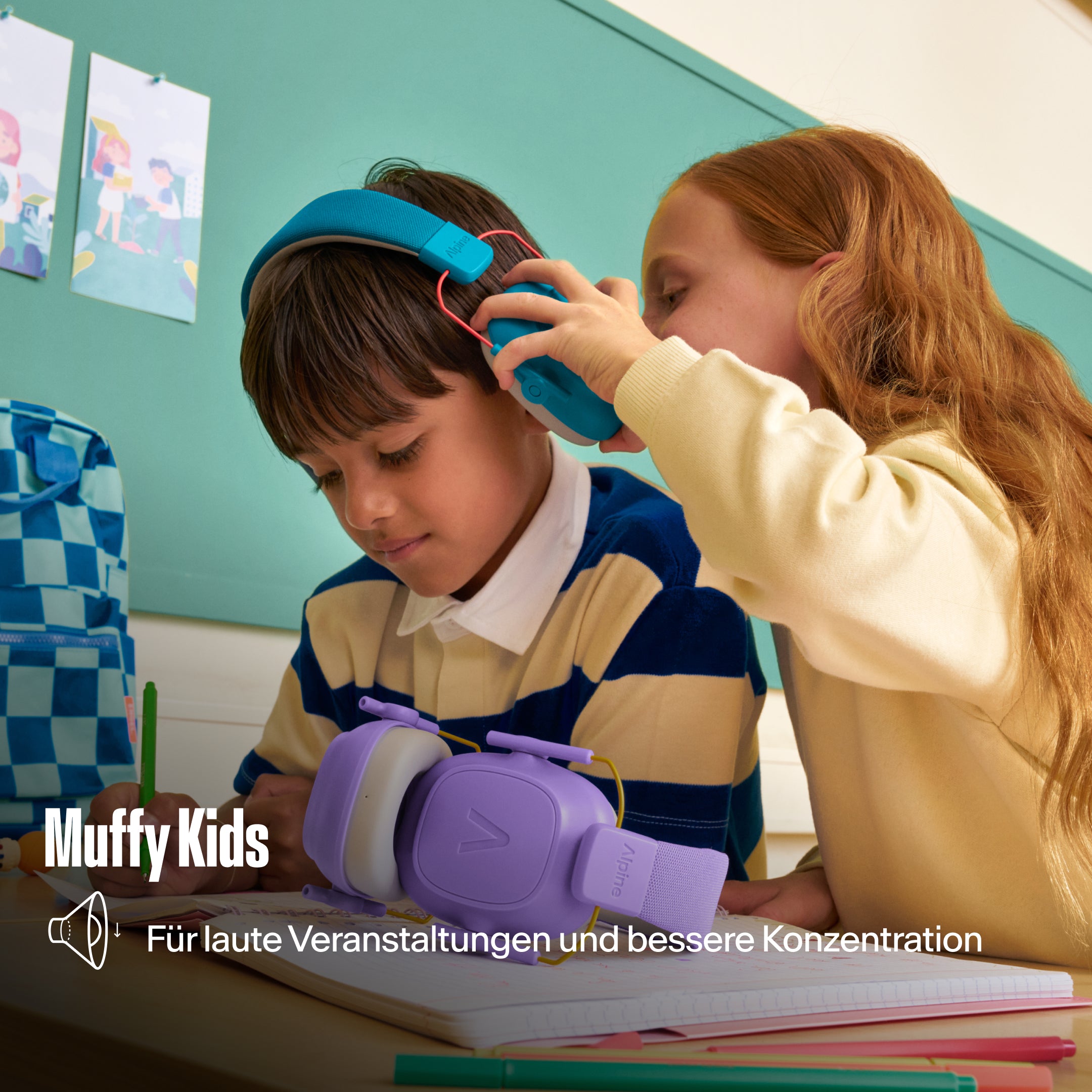 Alpine Muffy Kids Kapselgehörschutz, den ein Mädchen einem Jungen am Schultisch aufsetzt, mit dem Text &