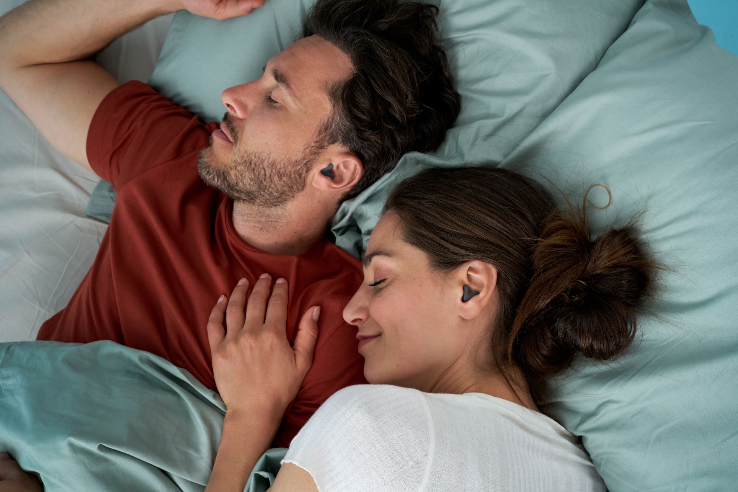 Young couple laying on the bed next to each other, both of them with Alpine Silence Black in their ears and smiling. The woman has her hand on the man&