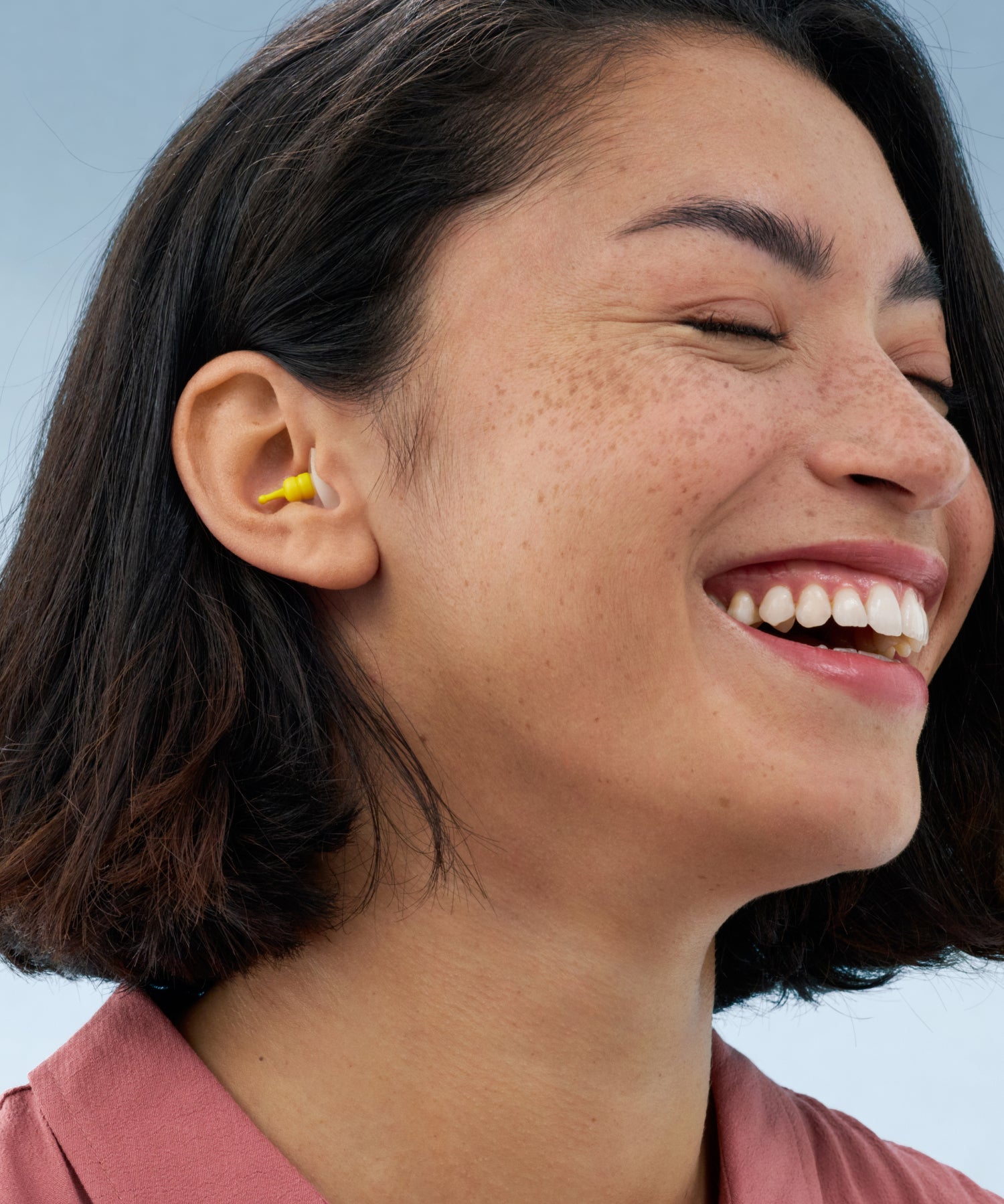 Alpine FlyFit earplug worn in the ear by a smiling woman with dark shoulder-length hair 